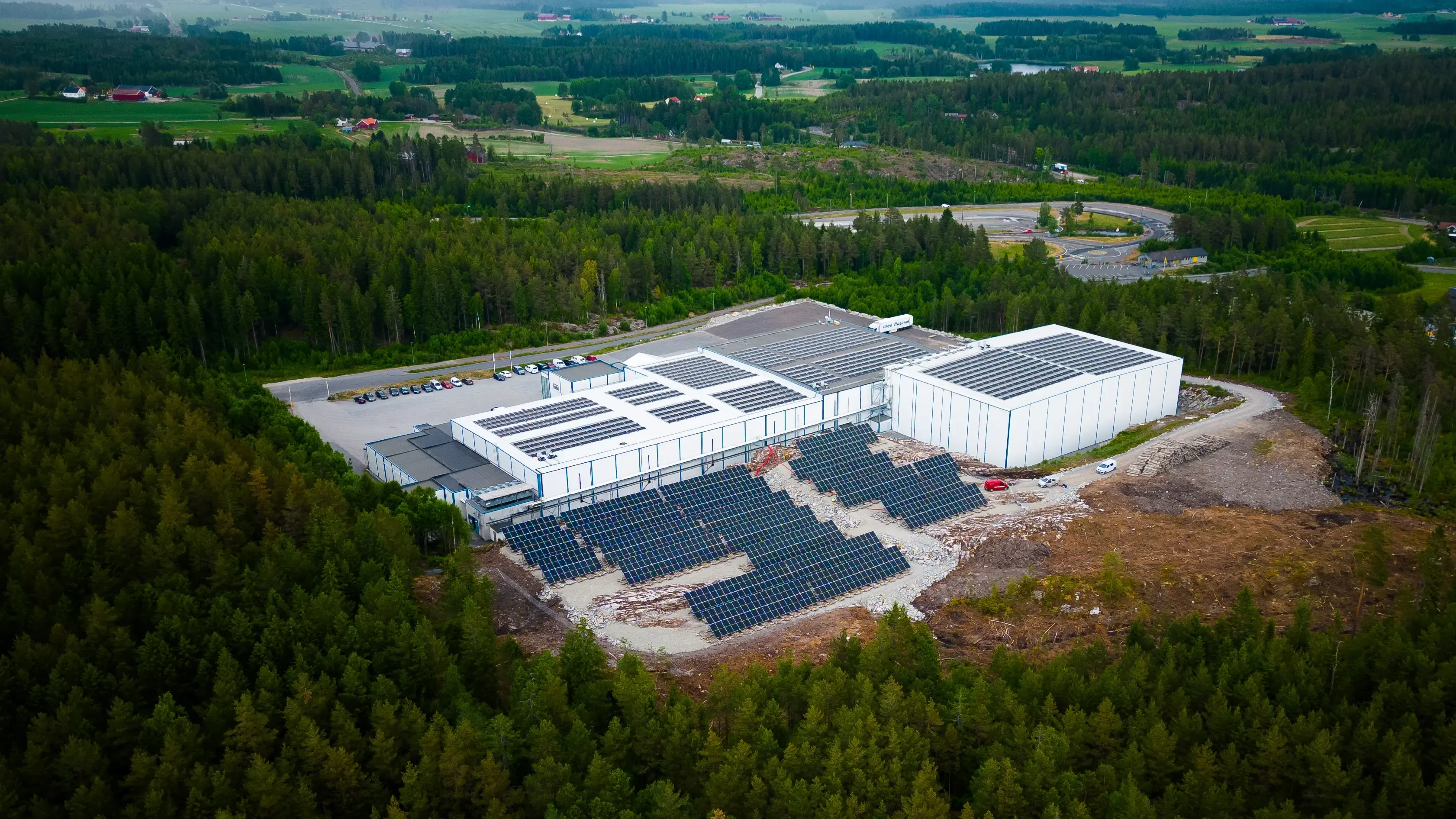 Aerial photograph of an industrial building in forest with solar panels on the roofs and ground-mounted rows on the cleared ground beside it